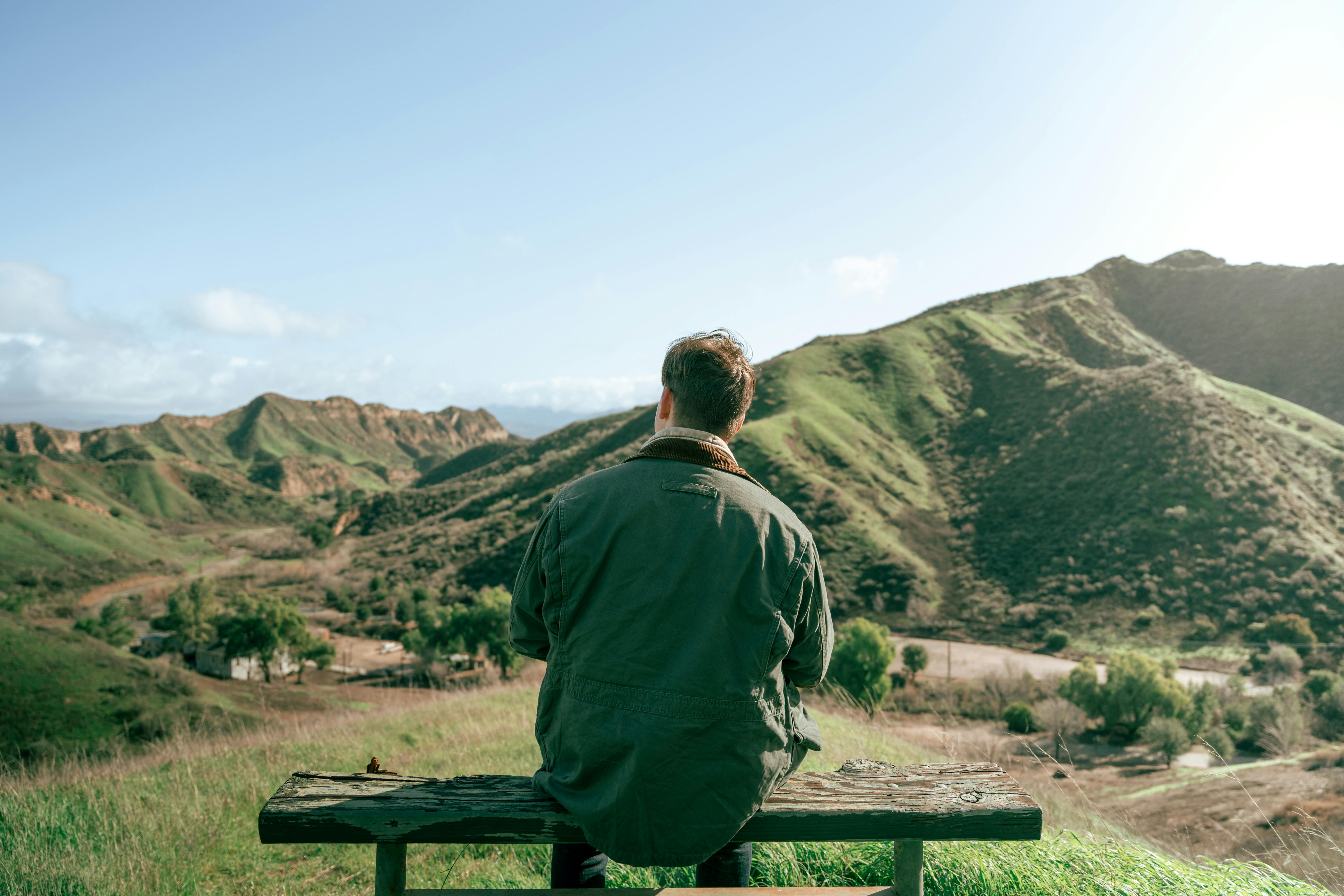 Man dressed in green jacket sits alone on bench atop a mountain of green hills.