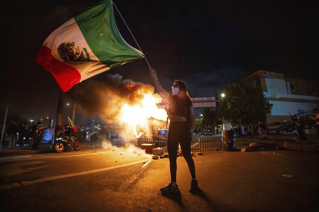 Woman waves the Mexican flag as flames erupt from a burning dumpster during a protest in downtown Los Angeles