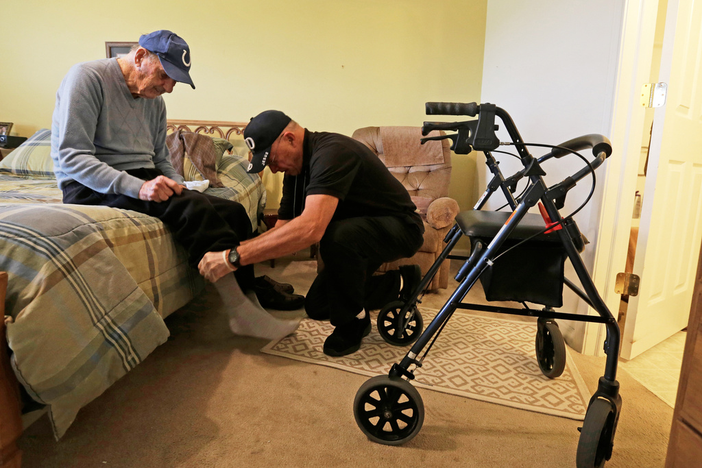Caregiver Warren Manchess helping Paul Gregoline with his shoes and socks, in Noblesville, Ind.