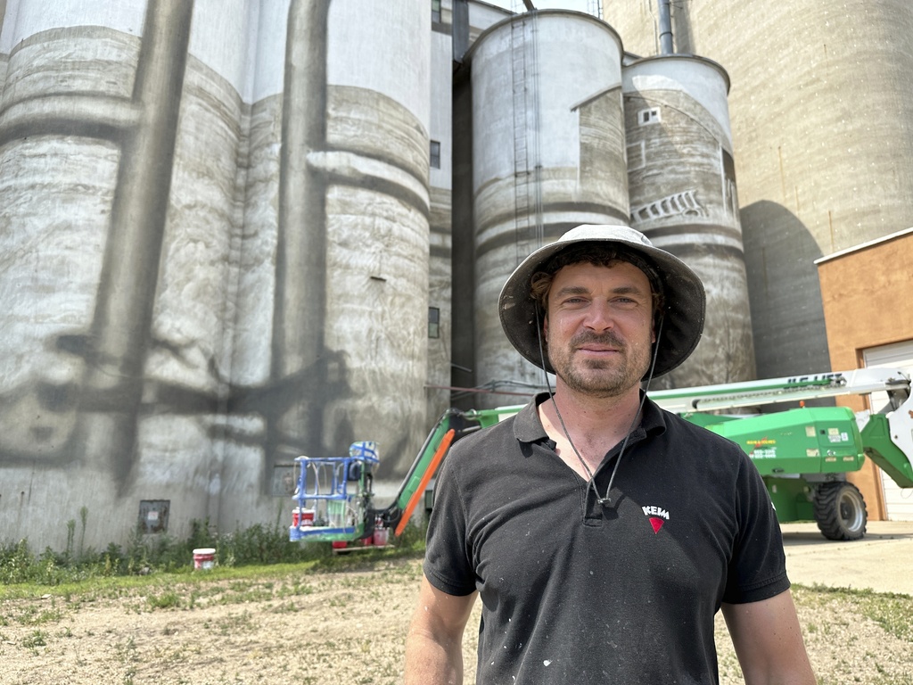 Artist Guido van Helten in front of the grain elevator and silos in Minot, N.D., where he is painting a large mural. Behind him is the boom lift he uses to ascend the structure. 