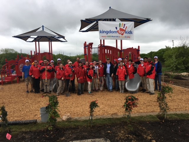 A large group of adults stand in front of a recently completed community service project, a playground.