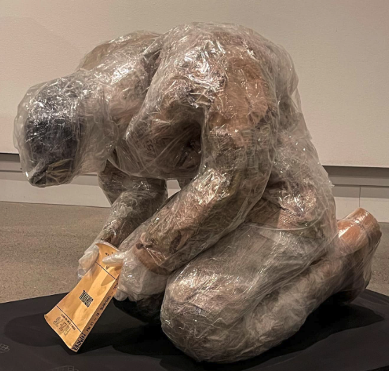 A sculpture of paper and plastic wrap of a man bent over, holding a survival guide