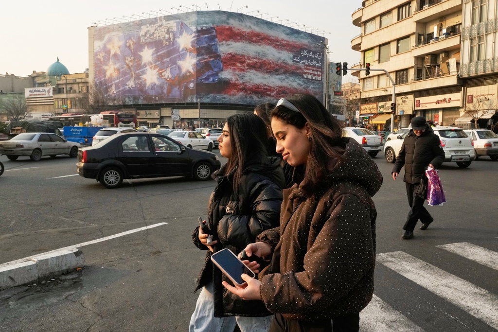 Two girls, not wearing the legally required headscarves, walk past a billboard depicting a damaged U.S. aircraft carrier with disabled fighter jets on its deck and a sign reading in Farsi and English, "If you sow the wind, you
