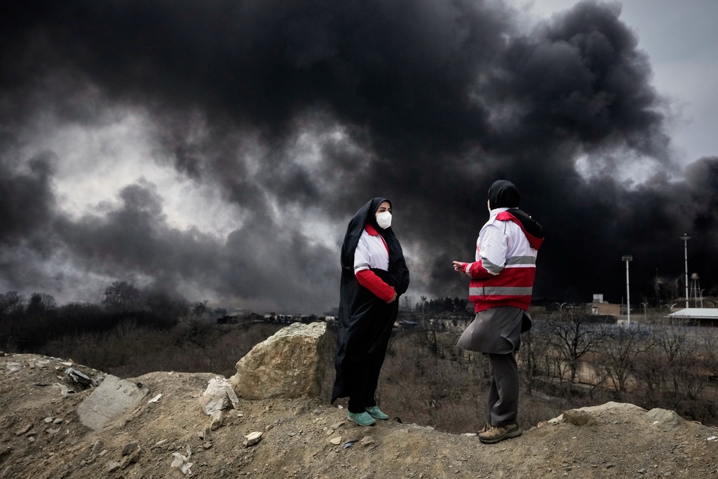 Two women from the Iranian Red Crescent Society stand as a thick plume of smoke from a U.S.-Israeli strike on an oil storage facility late Saturday rises in the sky in Tehran