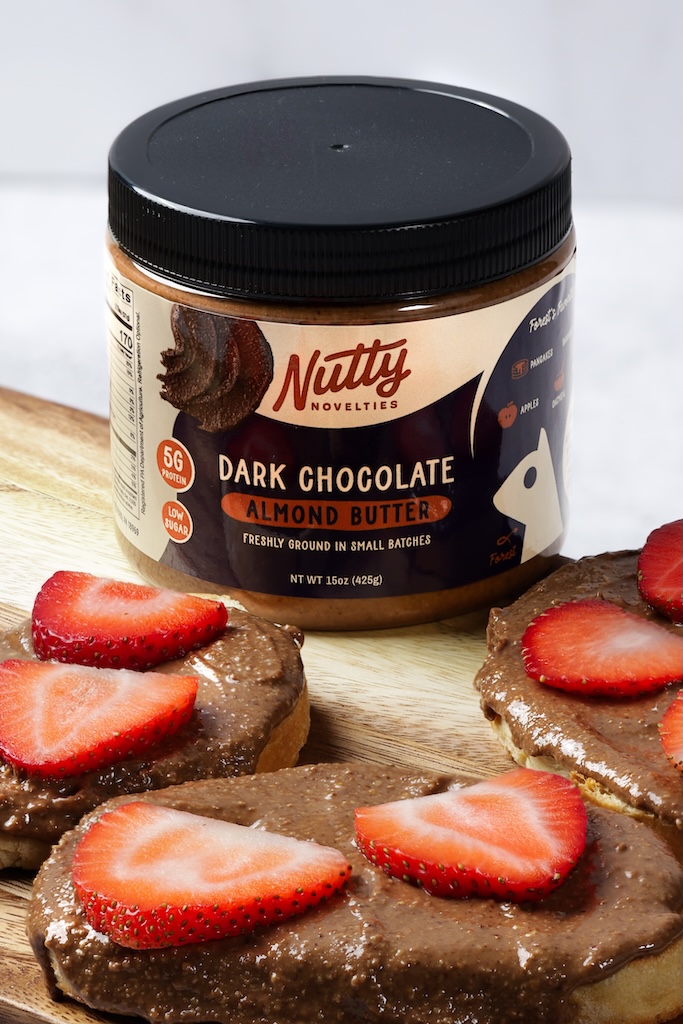 A jar of Nutty Novelties dark chocolate almond butter sits alongside toast topped with the spread and fresh strawberries. 