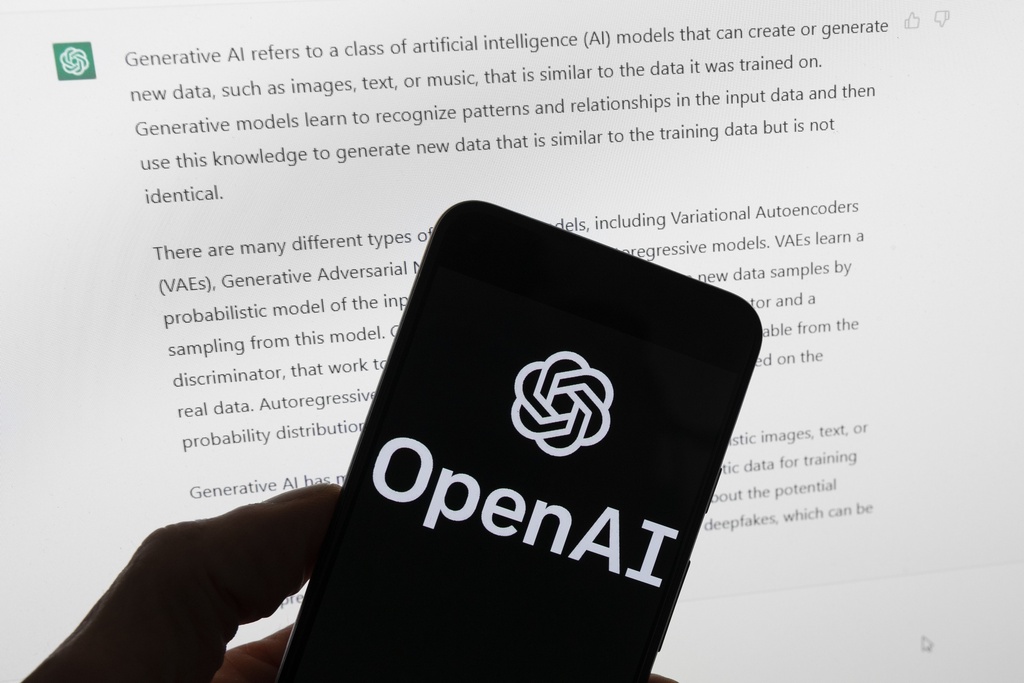 OpenAI logo is seen on a mobile phone in front of a computer screen displaying output from ChatGPT