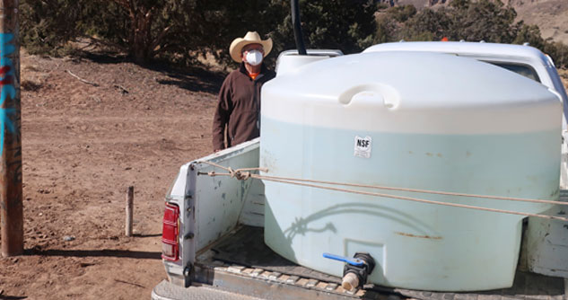 Man wearing a mask and cowboy hat filling the water drum in the back of his pickup truck