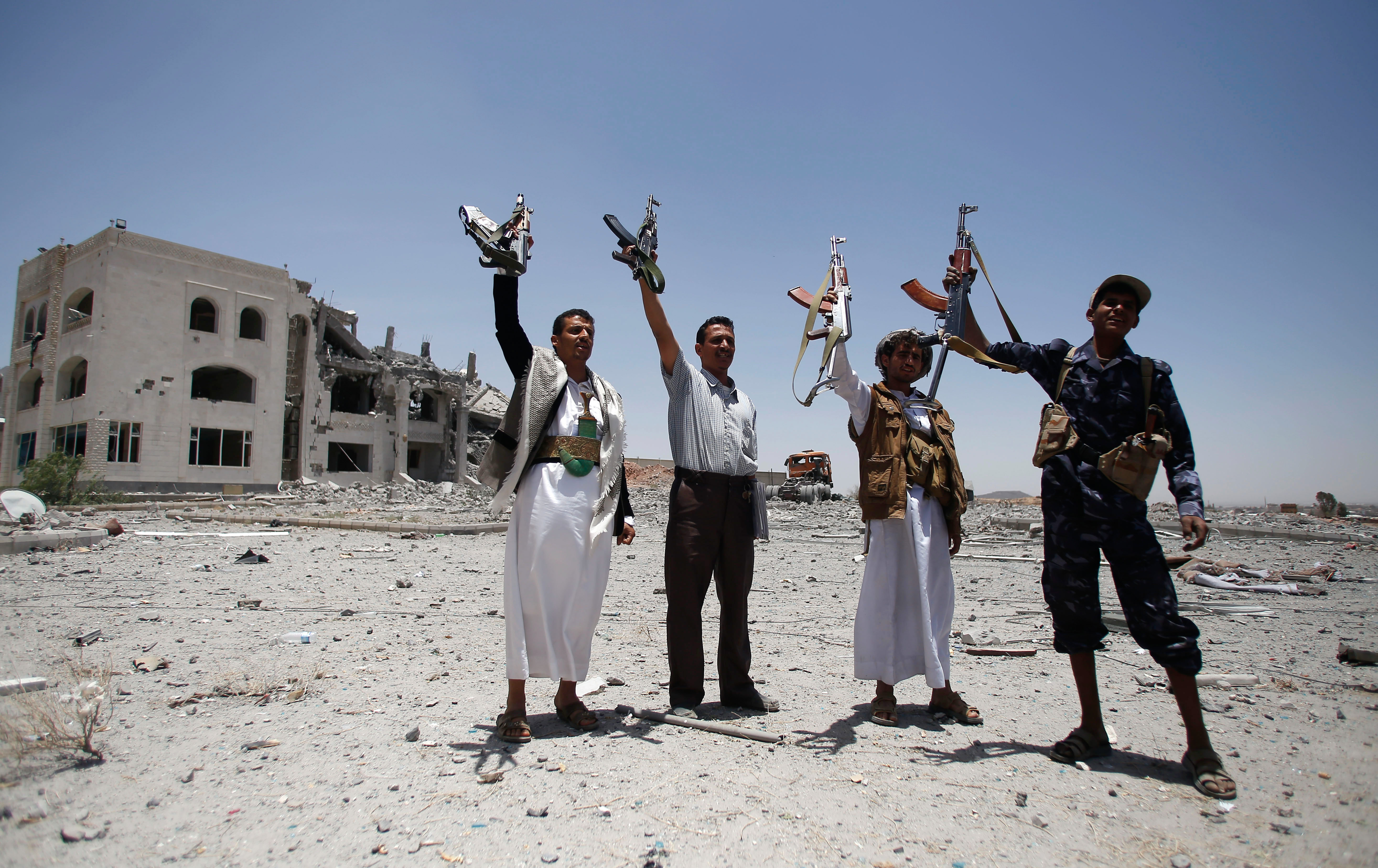 Four men holding up machine guns in front of a bombed-out building