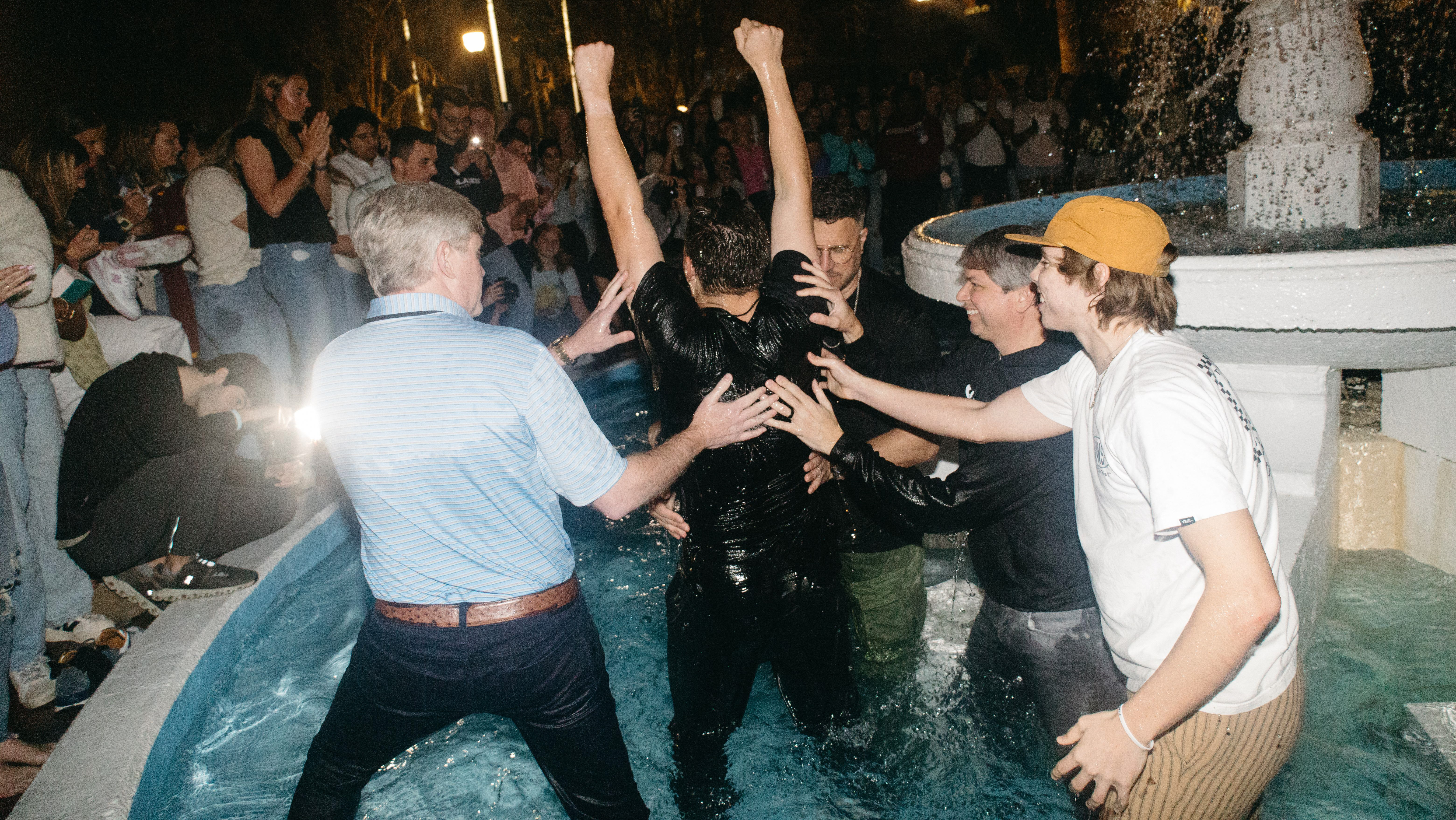 Four men helping to baptize a young man in a fountain
