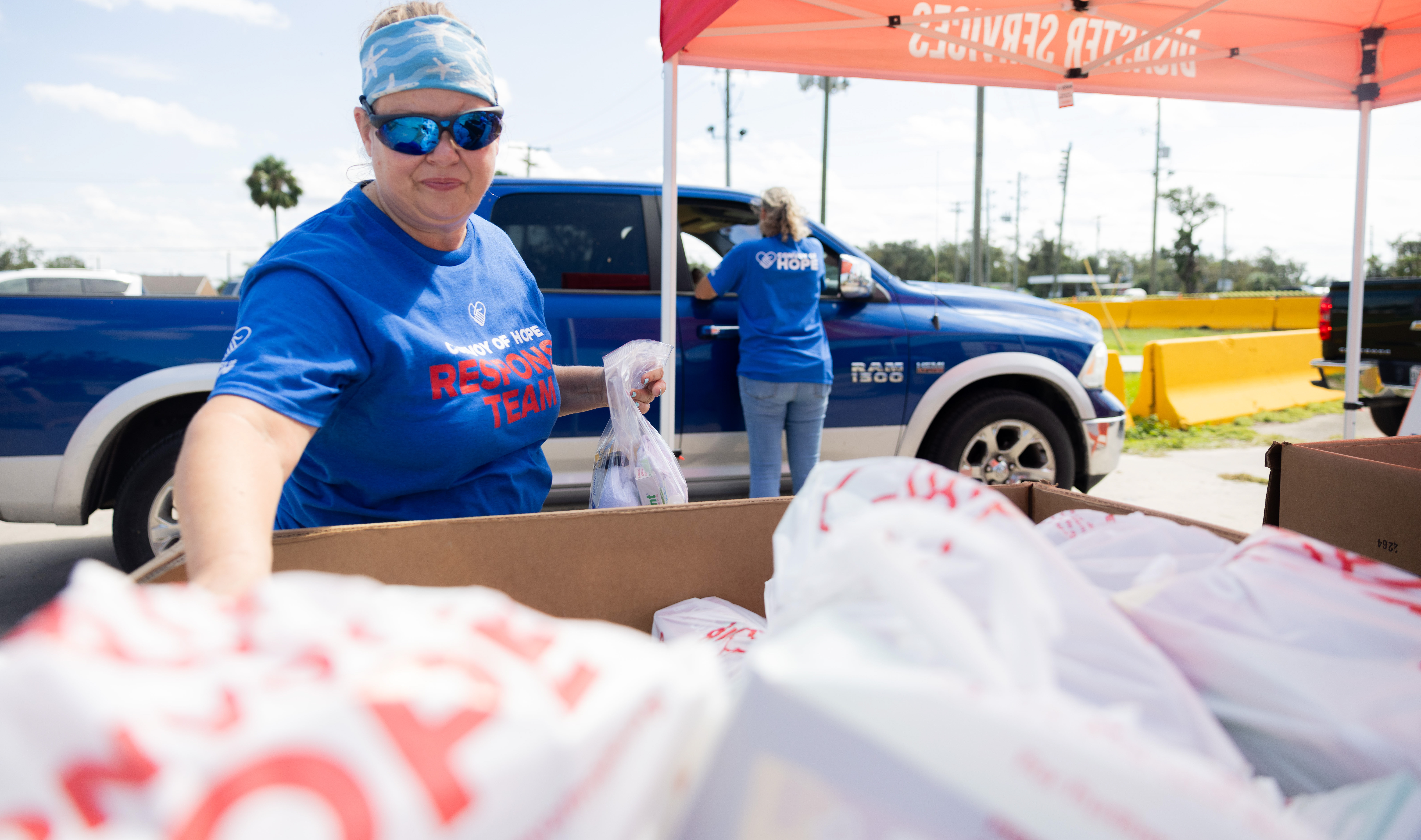 Woman reaching for a bag of groceries in the foreground, woman in the background bringing groceris to a truck background